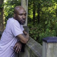 Middle-aged black man leaning on fence holding glasses.
