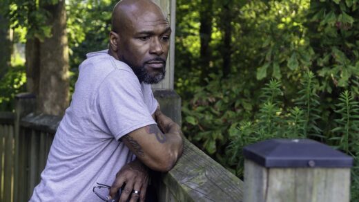 Middle-aged black man leaning on fence holding glasses.